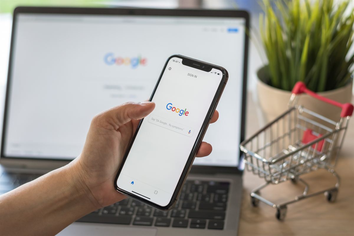 Hand hält ein Smartphone mit geöffneter Google-Suche vor einem Laptop