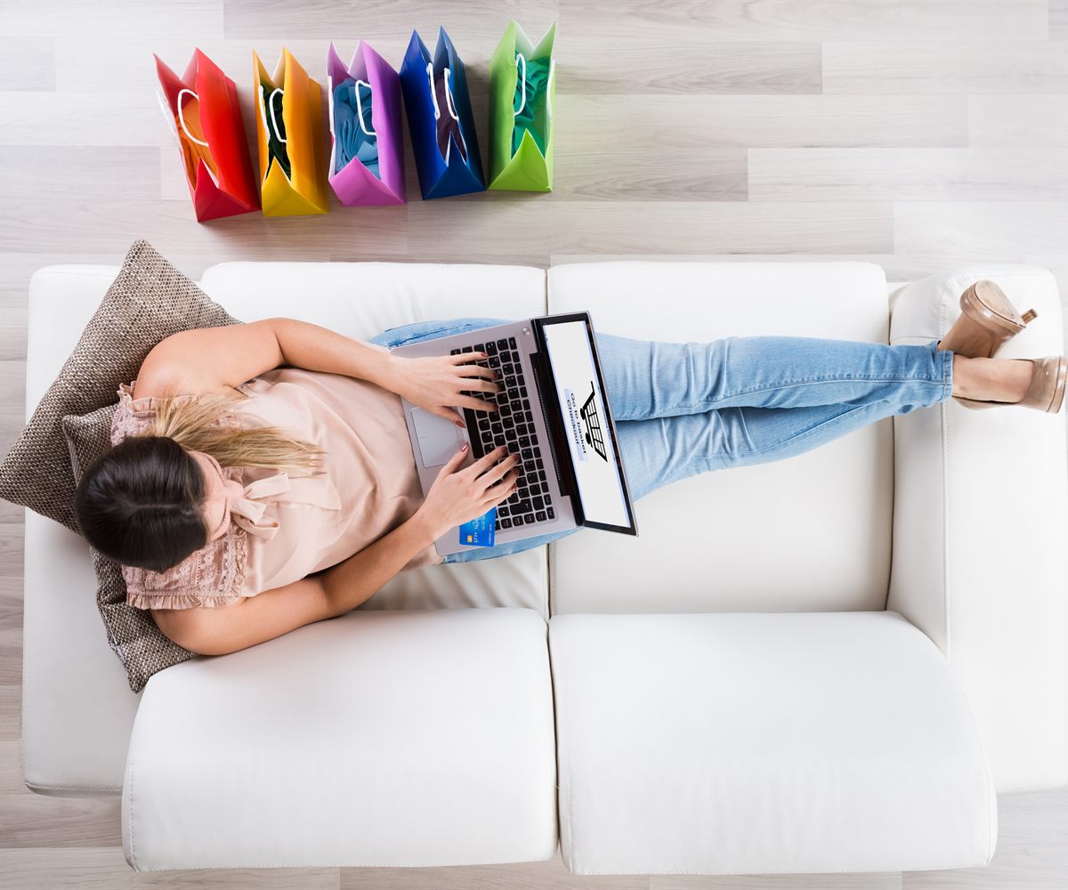 Frau liegt mit dem Laptop auf einem weißen Sofa und kauft online ein, daneben bunte Einkaufstüten