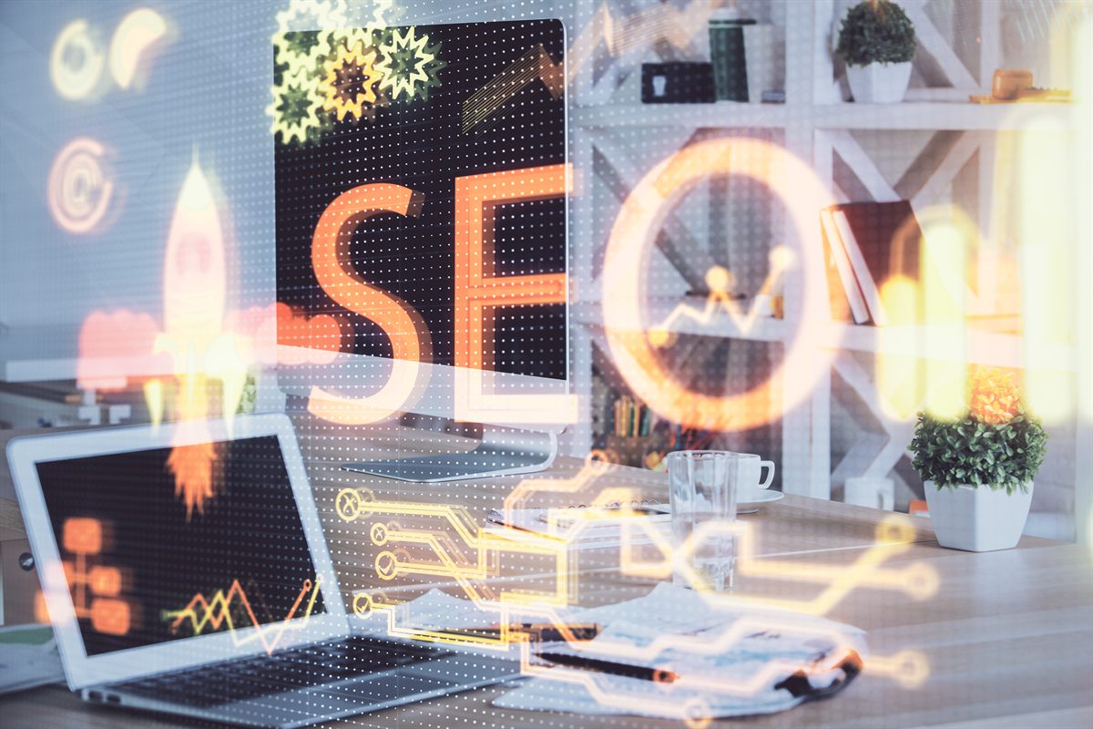 Arbeitsplatz mit Monitor und Laptop, davor eine leuchtende Grafik mit dem Schriftzug SEO, einer Rakete und Diagrammen