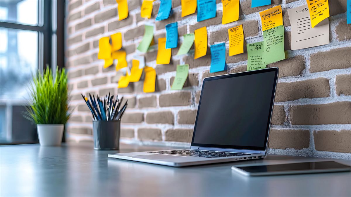 Laptop auf einem Schreibtisch vor einer Backsteinwand mit bunten Haftnotizen, daneben eine Pflanze und ein Stiftebecher
